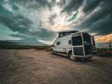 Experience outdoor travel with an RV rental! Modern pop-top camper van on a gravel road, mountains under dramatic skies.