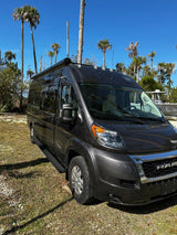 Dark grey Ram ProMaster camper van, ideal for an RV rental, parked on grass near palm trees under a blue sky.