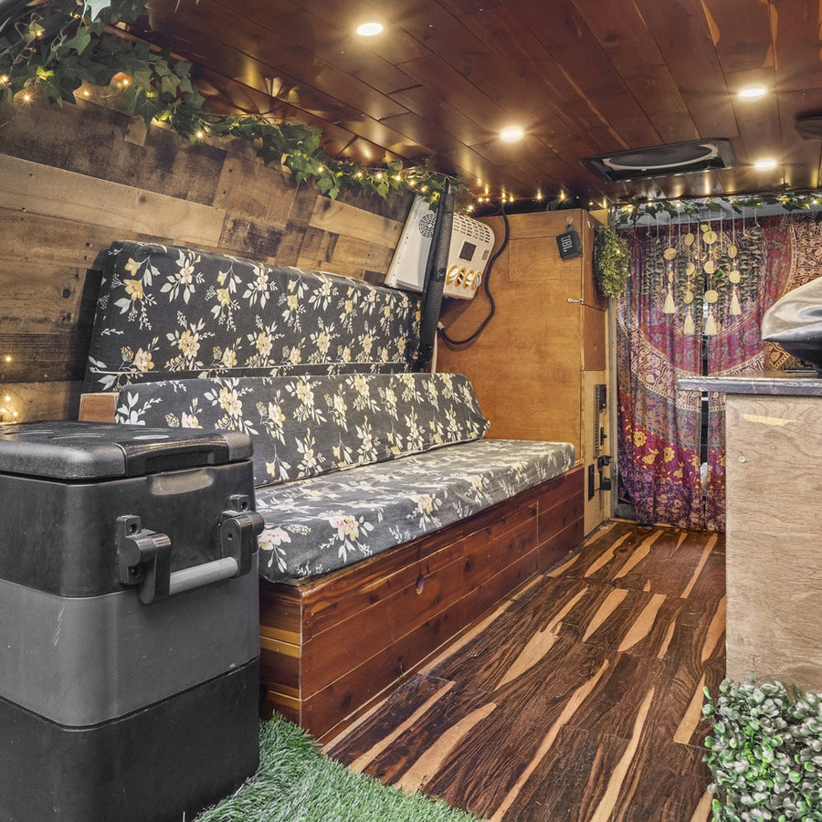 This RV rental's cozy interior features floral bench seating, wooden walls & ceiling, and ambient string lights.