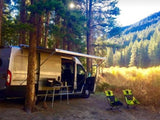 RV rental set up for camping in a sunny forest with mountains. A table, chairs, and awning create a cozy outdoor living space.