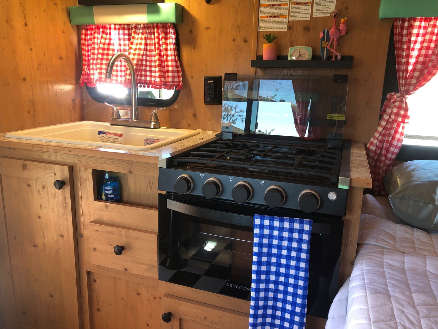 Cozy RV rental interior shows kitchen with sink, stove, wood cabinets, and red checkered curtains next to a sleeping area. — photo 9