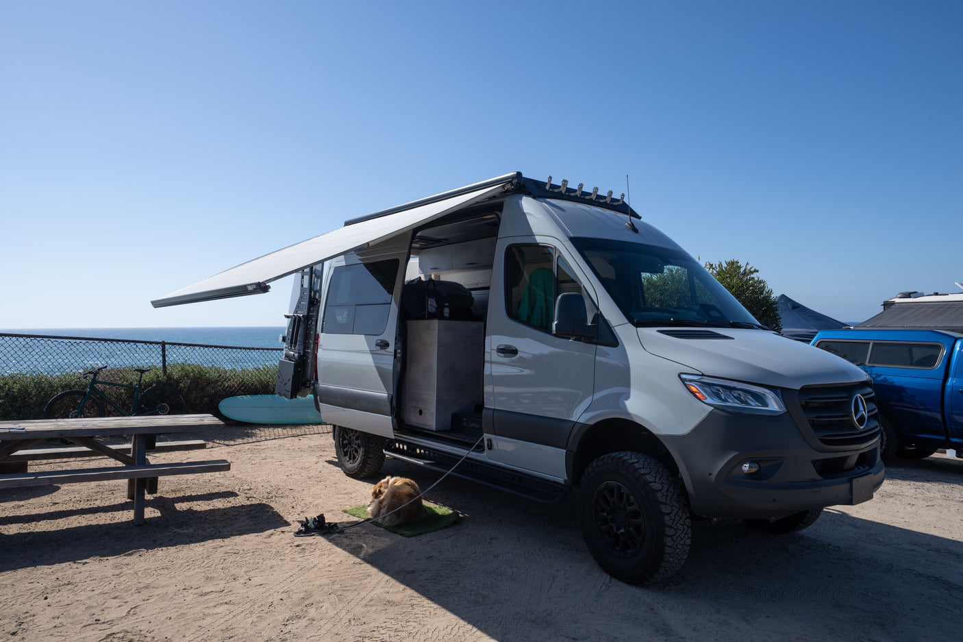 An RV rental Mercedes Sprinter van with extended awning parked near the ocean. A dog rests, a bike and surfboard are nearby.