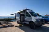 An RV rental Mercedes Sprinter van with extended awning parked near the ocean. A dog rests, a bike and surfboard are nearby.