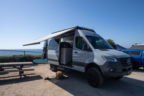 An RV rental Mercedes Sprinter van with extended awning parked near the ocean. A dog rests, a bike and surfboard are nearby.