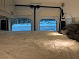Cozy rv rental interior: A bed with white sheets and gray bedding, offering a view of a snowy road and forest for winter travel.