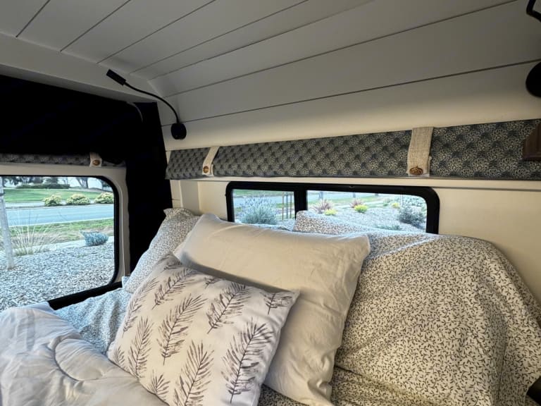 Cozy RV rental bedroom featuring soft bedding, a flexible reading light, and windows offering scenic views for comfortable travel.