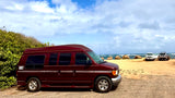 Maroon high-top van (RV rental) on a beach. Ocean, rocks & other vehicles visible under a cloudy sky.
