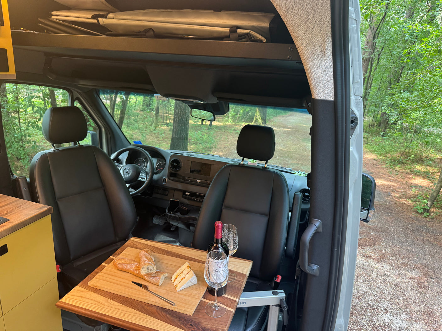 Cozy RV rental interior: a dining setup with wine, bread & cheese on a wooden table, overlooking a forest path from the van.