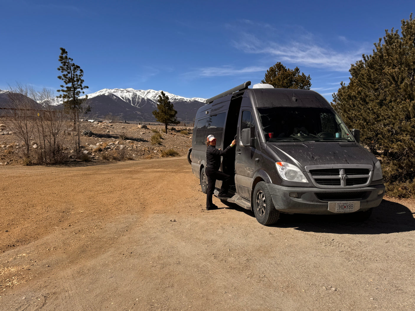 Woman steps into her RV rental camper van on a dirt path, set against snow-capped mountains for a mountain adventure.