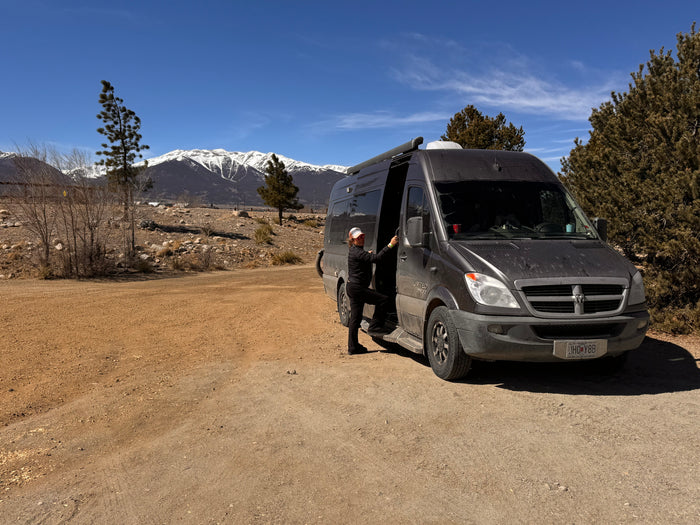 Woman steps into her RV rental camper van on a dirt path, set against snow-capped mountains for a mountain adventure.