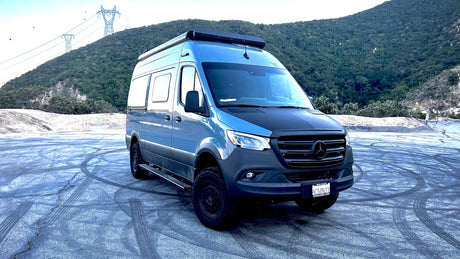 This rugged blue Mercedes-Benz Sprinter camper van, an ideal rv rental, is ready for adventure, parked before a forested mountain.