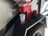 Red Keurig coffee maker and travel cups on an outdoor shelf of an RV rental, offering roadside convenience.