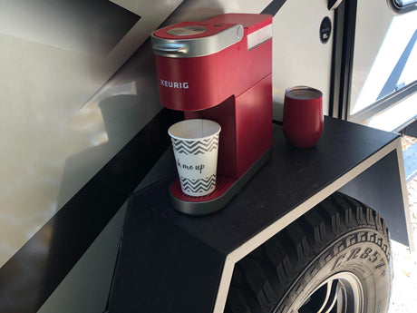 Red Keurig coffee maker and travel cups on an outdoor shelf of an RV rental, offering roadside convenience.