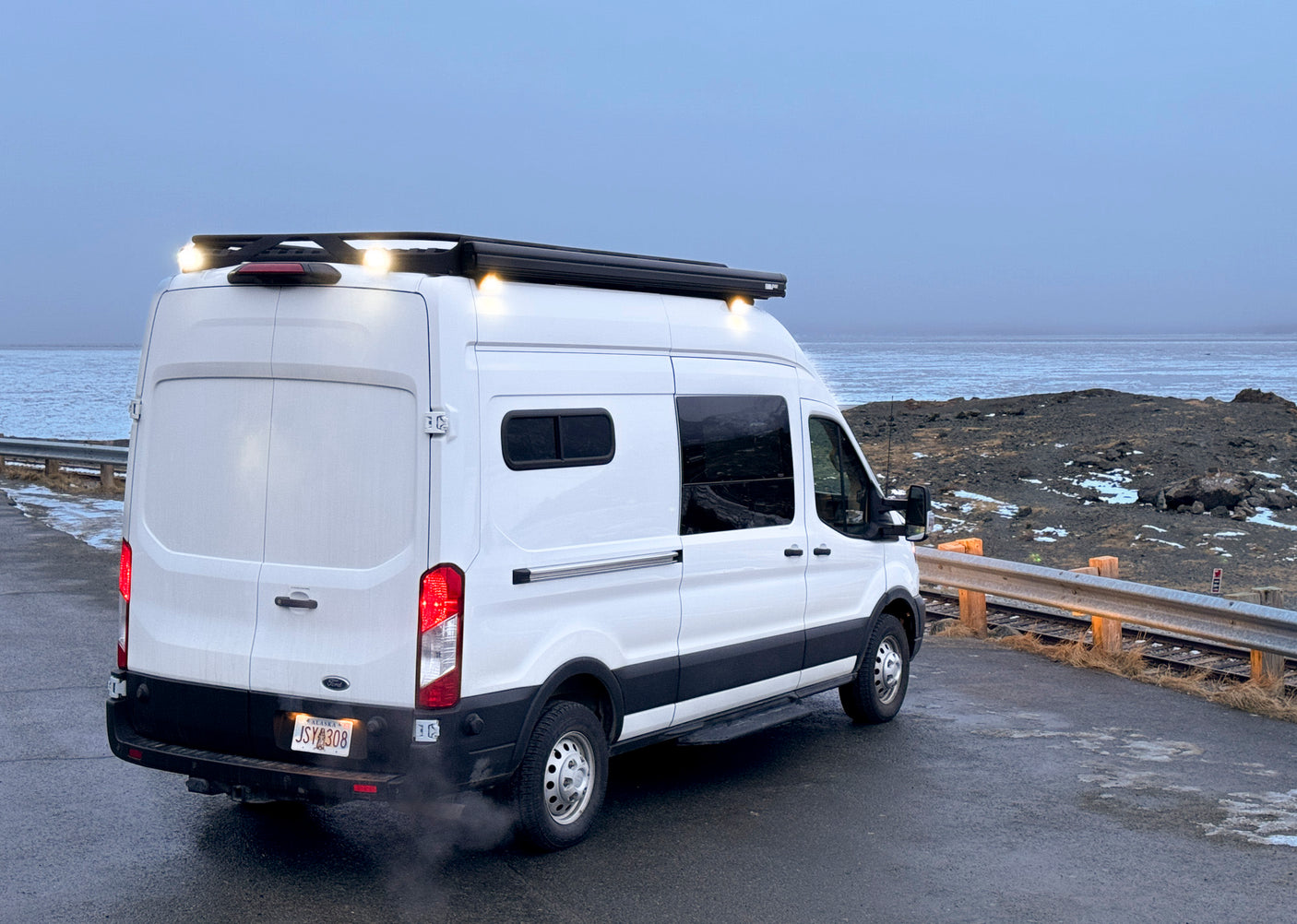 White Ford Transit RV rental camper van with roof lights, Alaskan plate, parked on a wet road by a grey sea & rocky coast.