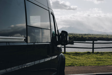 This dark RV rental is parked by a serene lake, offering stunning views of distant mountains beyond a wooden fence.