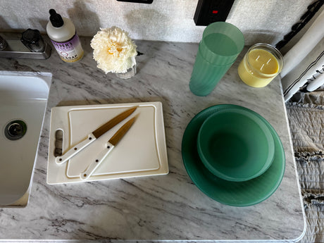 Compact RV rental kitchen counter with sink, soap, knives on board, and green plates/glass, ready for adventure.