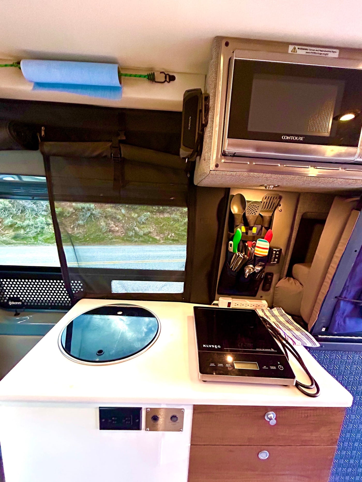 This RV rental's compact kitchen features a covered sink, portable cooktop, microwave, and utensils, overlooking a road. — photo 15