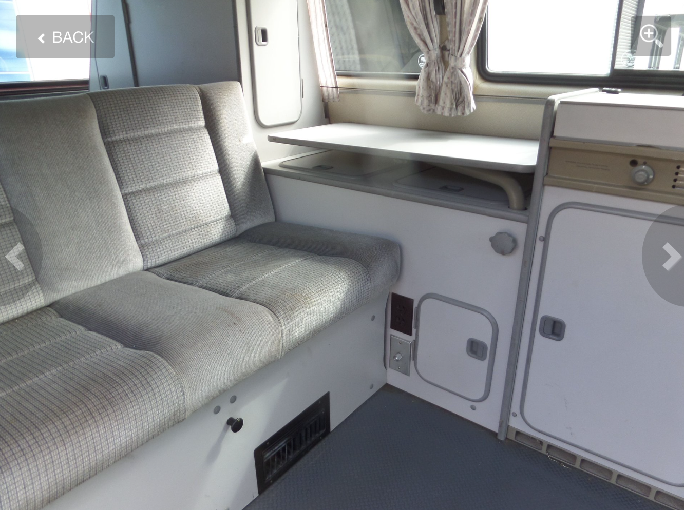 Interior of an RV rental: view of a gray patterned bench seat, fold-out table, and beige kitchen unit with storage and controls.
