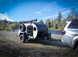 An RV rental: a compact, black and white teardrop camper is hitched to a silver SUV, parked by a scenic river and forest.