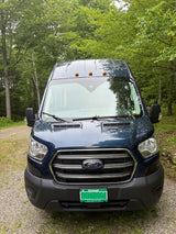 This blue Ford Transit van RV rental is parked on a gravel path, surrounded by lush green trees.