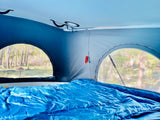 RV rental interior with cozy blue bedding, offering scenic views of a forest campground from pop-top windows.