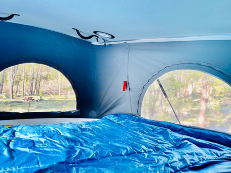 RV rental interior with cozy blue bedding, offering scenic views of a forest campground from pop-top windows.