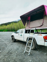 White pickup with a vibrant pink roof-top tent ready for an RV rental adventure, parked on gravel by mountains and forest.