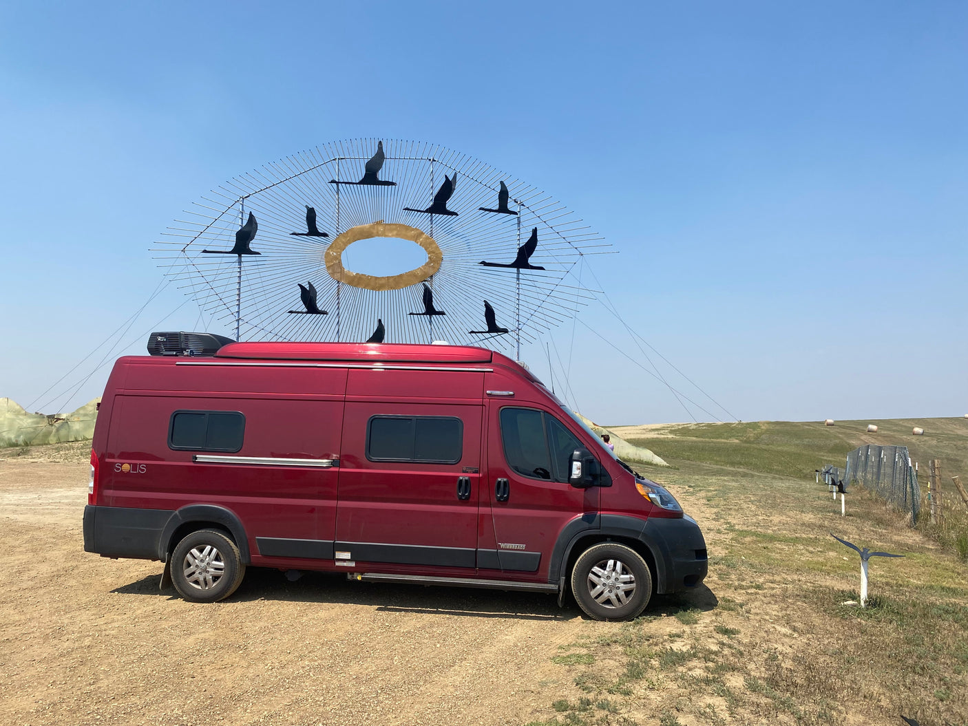 This red RV rental is parked by a captivating outdoor sculpture of silhouetted birds in flight around a golden ring under a clear sky.