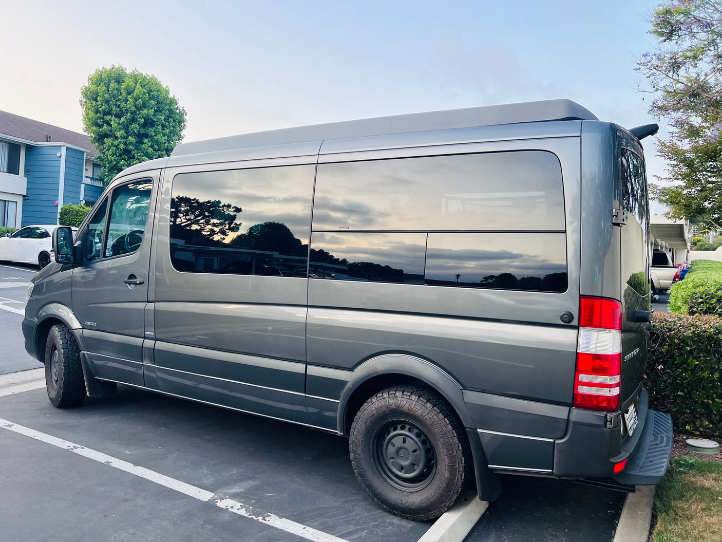 Dark gray Mercedes Sprinter rv rental camper van, with tinted windows & all-terrain tires, parked outdoors.