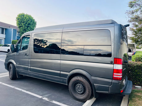 Dark gray Mercedes Sprinter rv rental camper van, with tinted windows & all-terrain tires, parked outdoors.