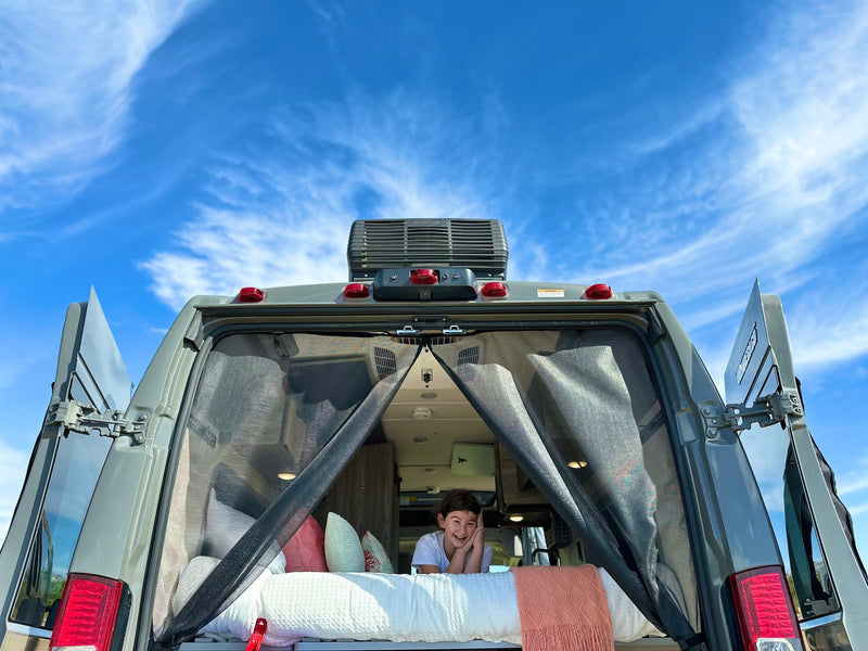  ready for adventure overlooking a dirt road.|Inside a comfy RV rental: a bed and a screened window with a beautiful view of a calm bay and sandy beach.|A smiling woman enjoys the view from the pop-top of an RV rental — photo 11