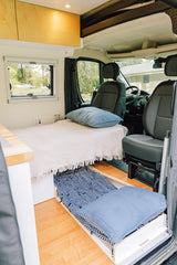 Inside a modern rv rental: a cozy bed, pull-out floor cushion, light wood cabinets, and clear view to the front cabin.