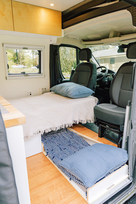 Inside a modern rv rental: a cozy bed, pull-out floor cushion, light wood cabinets, and clear view to the front cabin.