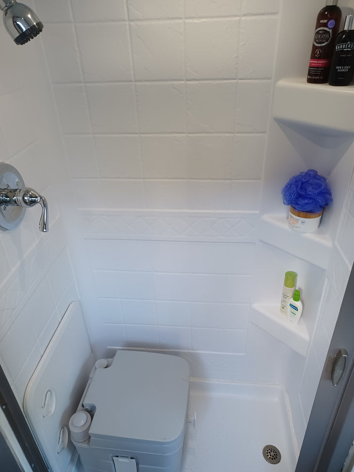 Inside an RV rental shower, showing a toilet, shower head, and toiletries: shampoo, body butter, Cetaphil lotion, and a loofah.