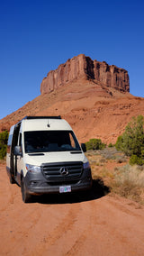 Scenic RV rental adventure: A white Mercedes Sprinter campervan on a red dirt road with massive red rock formations under a blue sky.