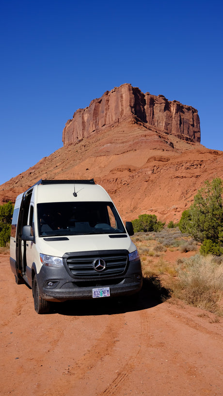 Scenic RV rental adventure: A white Mercedes Sprinter campervan on a red dirt road with massive red rock formations under a blue sky.
