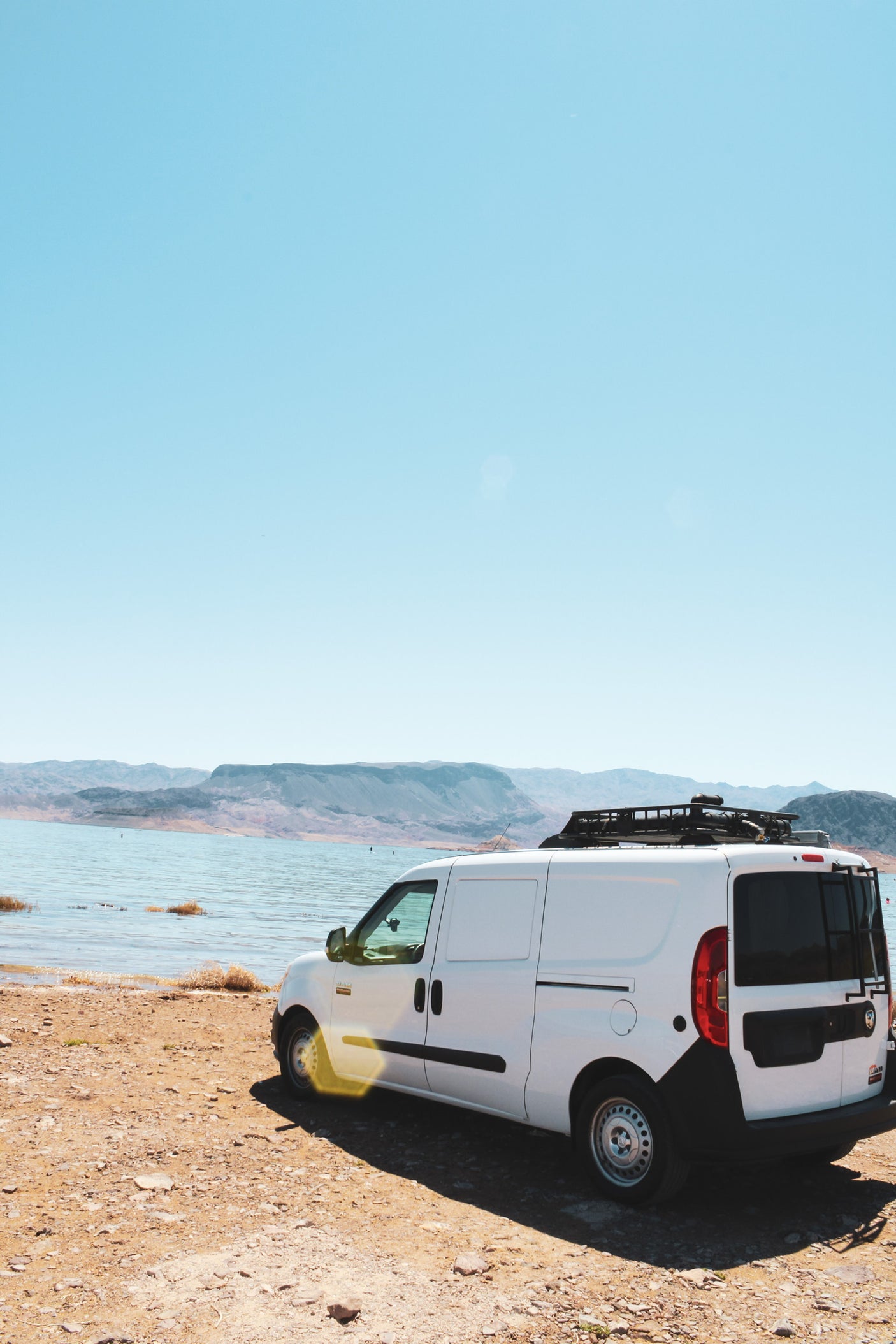 Adventure awaits! White RV rental campervan parked lakeside with mountains under a clear sky.
