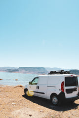 Adventure awaits! White RV rental campervan parked lakeside with mountains under a clear sky.
