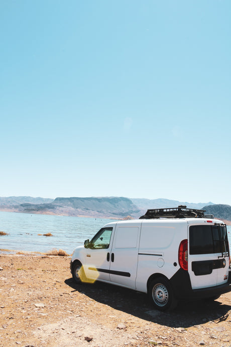 Adventure awaits! White RV rental campervan parked lakeside with mountains under a clear sky.