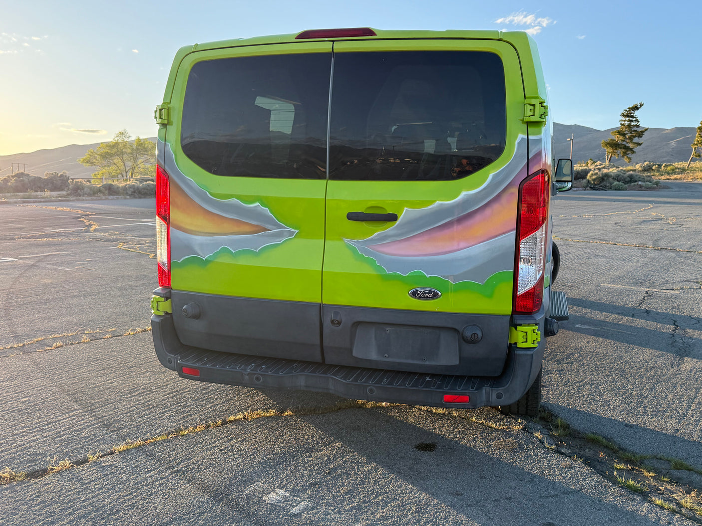 Bright green custom Ford Transit van, likely an RV rental, with a torn-look paint job, parked on asphalt at sunset with hills behind.