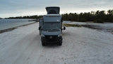 Grey RV rental on a sandy beach with pop-top extended and awnings deployed, facing the viewer with ocean and palms behind.