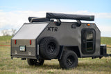 A rugged dark gray XTR off-road camper trailer in a grassy field. An ideal RV rental for backcountry adventures.