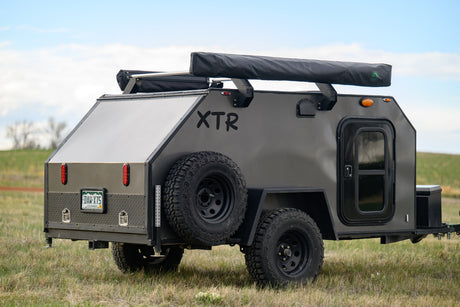 A rugged dark gray XTR off-road camper trailer in a grassy field. An ideal RV rental for backcountry adventures.