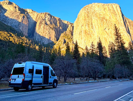 An RV rental campervan is parked by a road under immense, golden, sunlit mountains and a clear blue sky. Trees line the base.