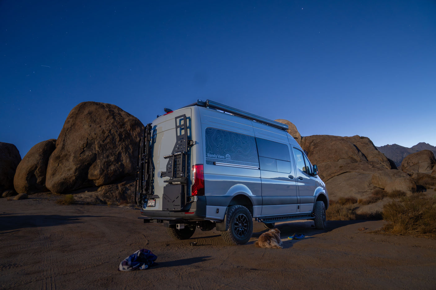 A grey Mercedes Sprinter RV rental campervan is parked at night in a rocky desert with a sleeping dog, under a starry sky.
