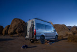 A grey Mercedes Sprinter RV rental campervan is parked at night in a rocky desert with a sleeping dog, under a starry sky.