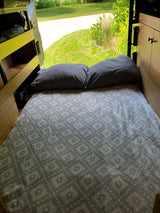 Comfortable RV rental bed with grey pillows & patterned blanket, looking out to a sunny green yard. Ideal for road trip rest.