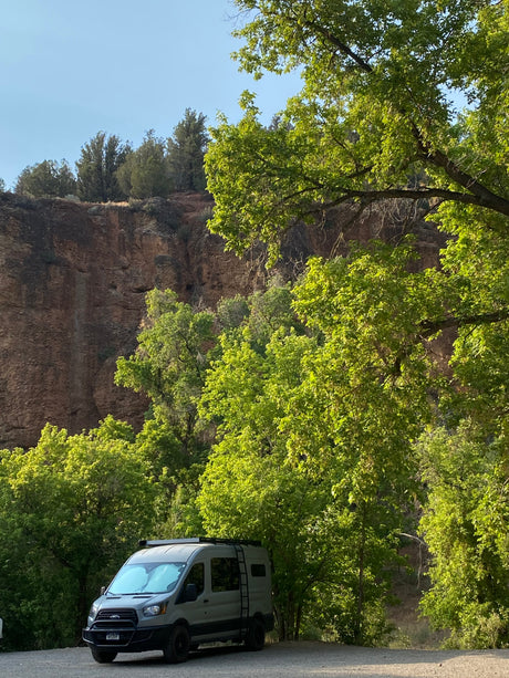 Explore nature with an RV rental! Grey camper van nestled among green trees, overlooking a majestic cliff.