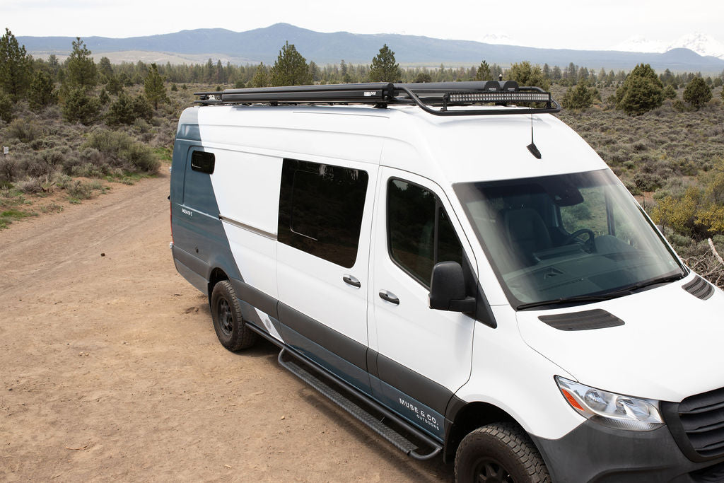 White & grey Sprinter camper van for RV rental, featuring a roof rack and LED light bar, parked on a dirt road in the mountains.