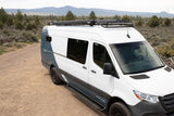 White & grey Sprinter camper van for RV rental, featuring a roof rack and LED light bar, parked on a dirt road in the mountains.